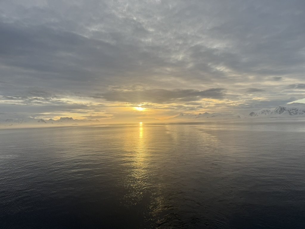 Sun breaking through cloud over open water scattered with ice, mountains on the horizon
