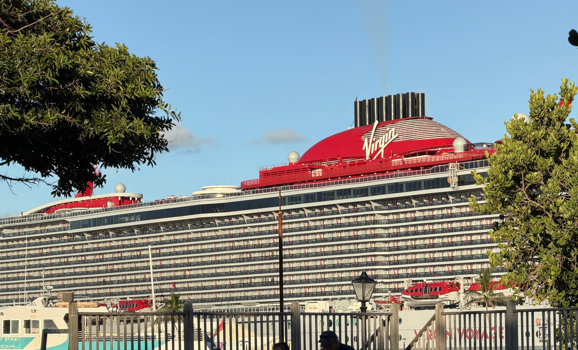 Virgin Voyages ship Valiant Lady, with its red funnel, docked behind trees at the Royal Naval Dockyard in Bermuda