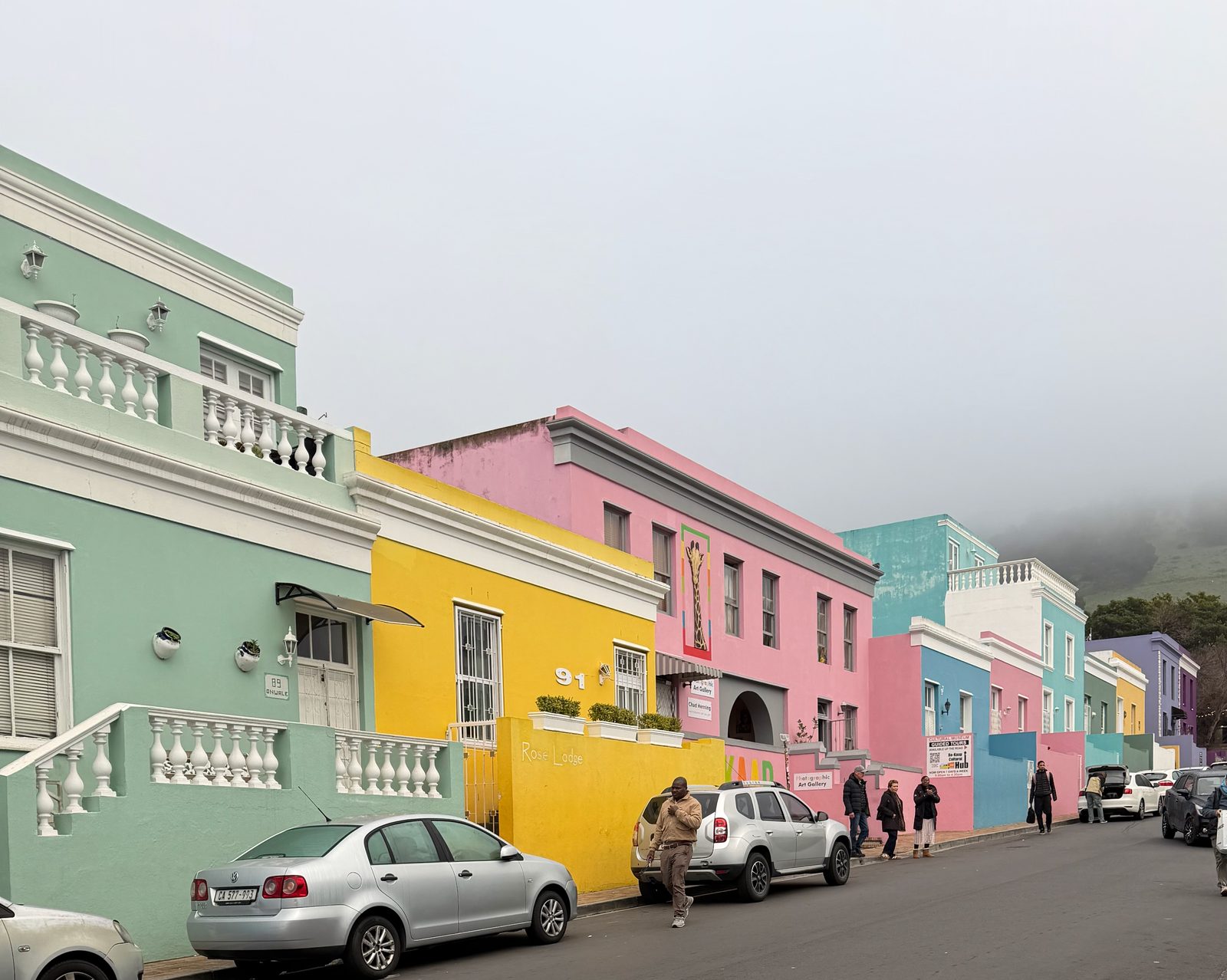 A steep street of brightly painted houses in pink, green and yellow under low cloud