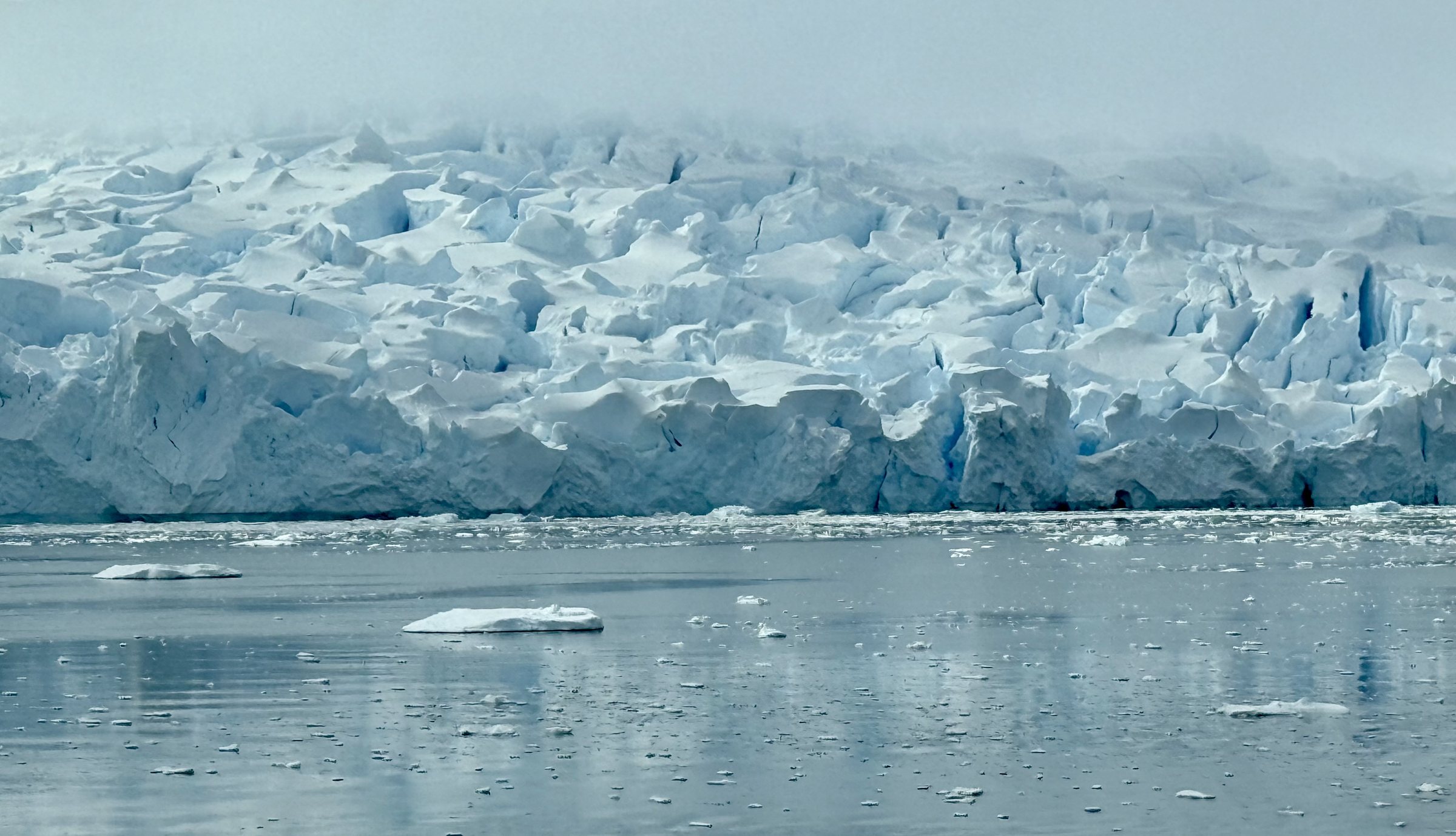 A long fractured glacier wall in soft light, brash ice scattered on the water below