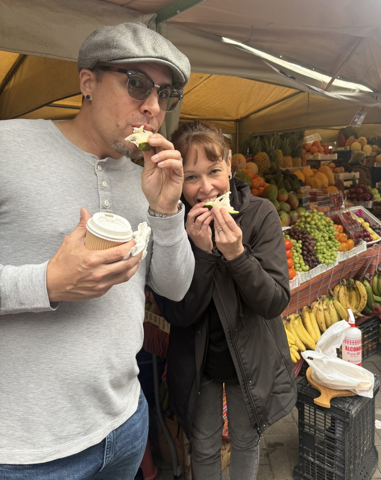 Two travellers tasting fresh fruit at a covered market stall stacked with tropical produce