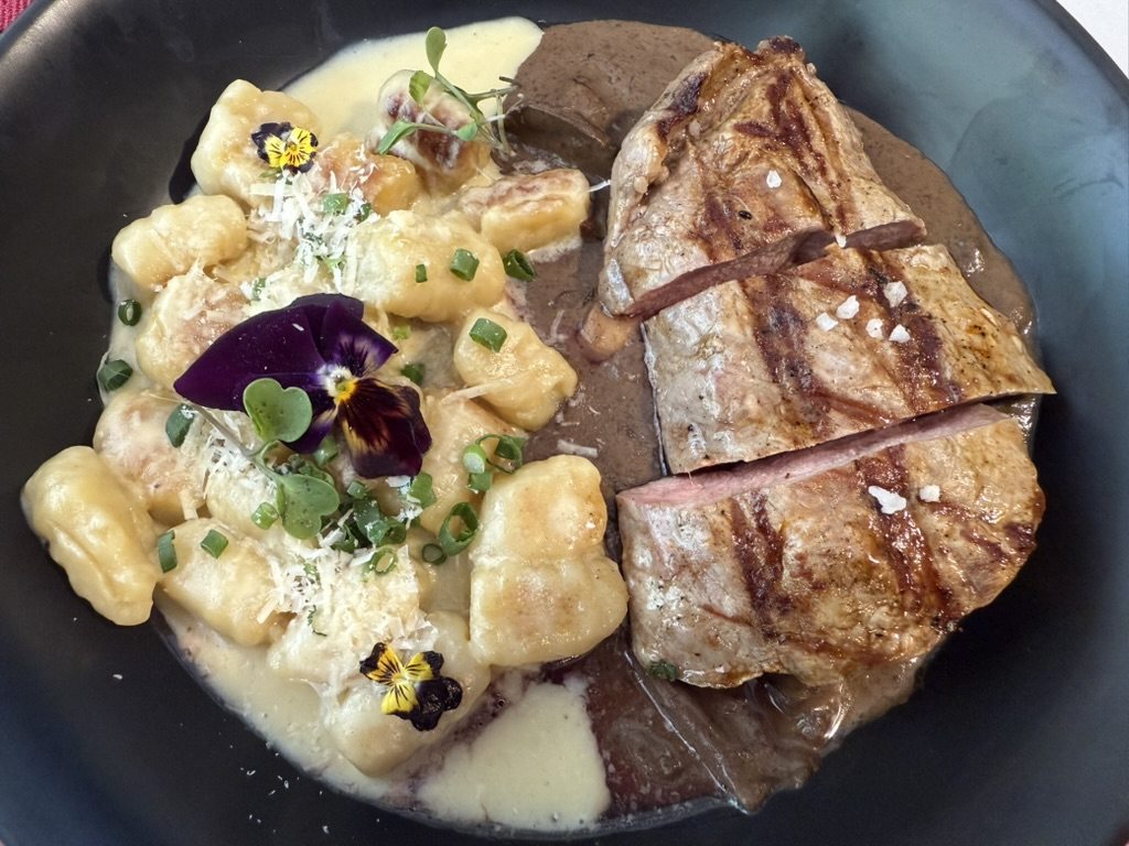 A plated dish of gnocchi in cream sauce with grilled meat and edible flowers