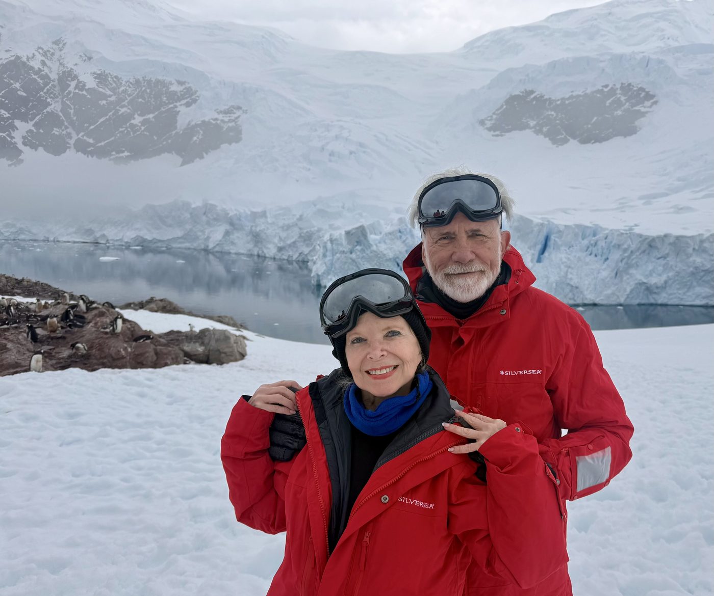Dana Wallace Cash and her husband in red expedition parkas, glacier and penguin colony behind them