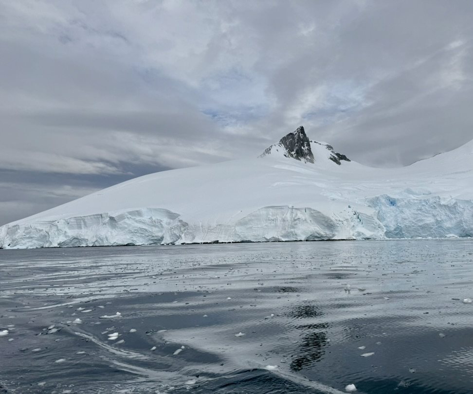 A dark rock peak rising above a smooth snow dome, dark water in the foreground