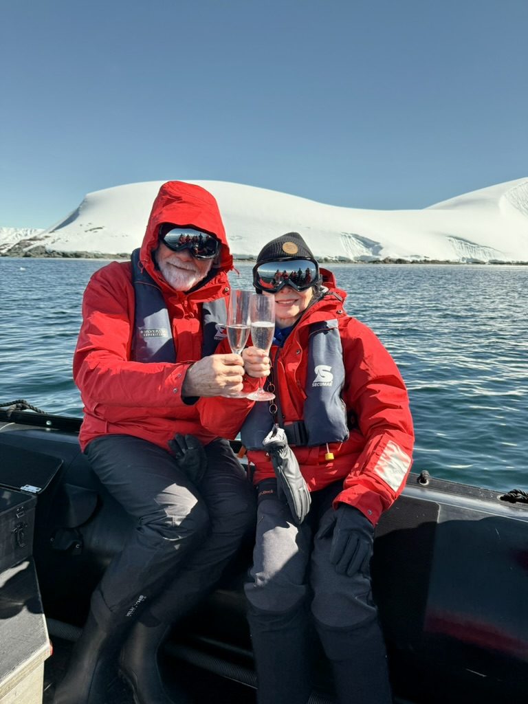 Two travellers in red expedition parkas raising glasses in a Zodiac, snow-covered island behind